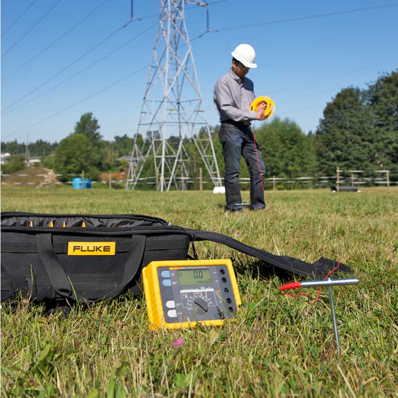 The FLUKE 1625-2 earth tester being used for ground testing near a power transmission line, showing its application in utility maintenance.