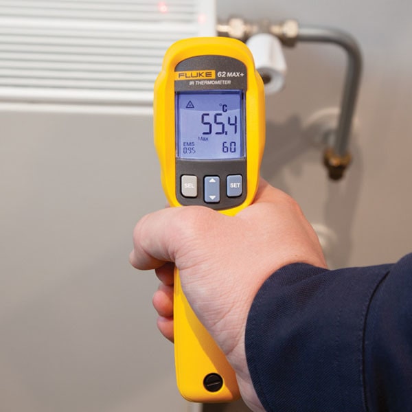 A maintenance technician holding the FLUKE 62 Max+ non-contact pyrometer and aiming the dual rotating lasers at an HVAC air conditioning refrigerant pipe line, showing 55.4°C temperature with a peak value of 60°C for AC troubleshooting and efficiency auditing.