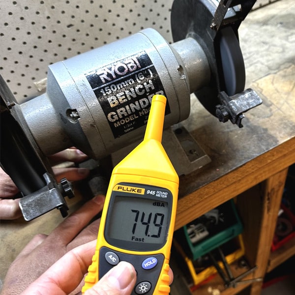 A technician monitoring industrial noise levels with a Fluke 945 sound meter, showing a 74.9 dBA reading with a bench grinder operating in the workshop.