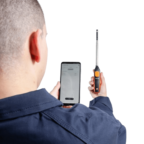 An HVAC technician holds the testo 405i hot-wire anemometer while monitoring real-time airflow data on the testo Smart App.