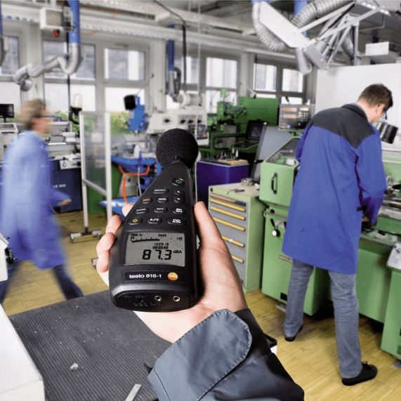 An industrial engineer measures high noise levels in a workshop with the TESTO 816-1 sound meter, which displays 87.3 dBA, ensuring OSHA compliance and worker safety.