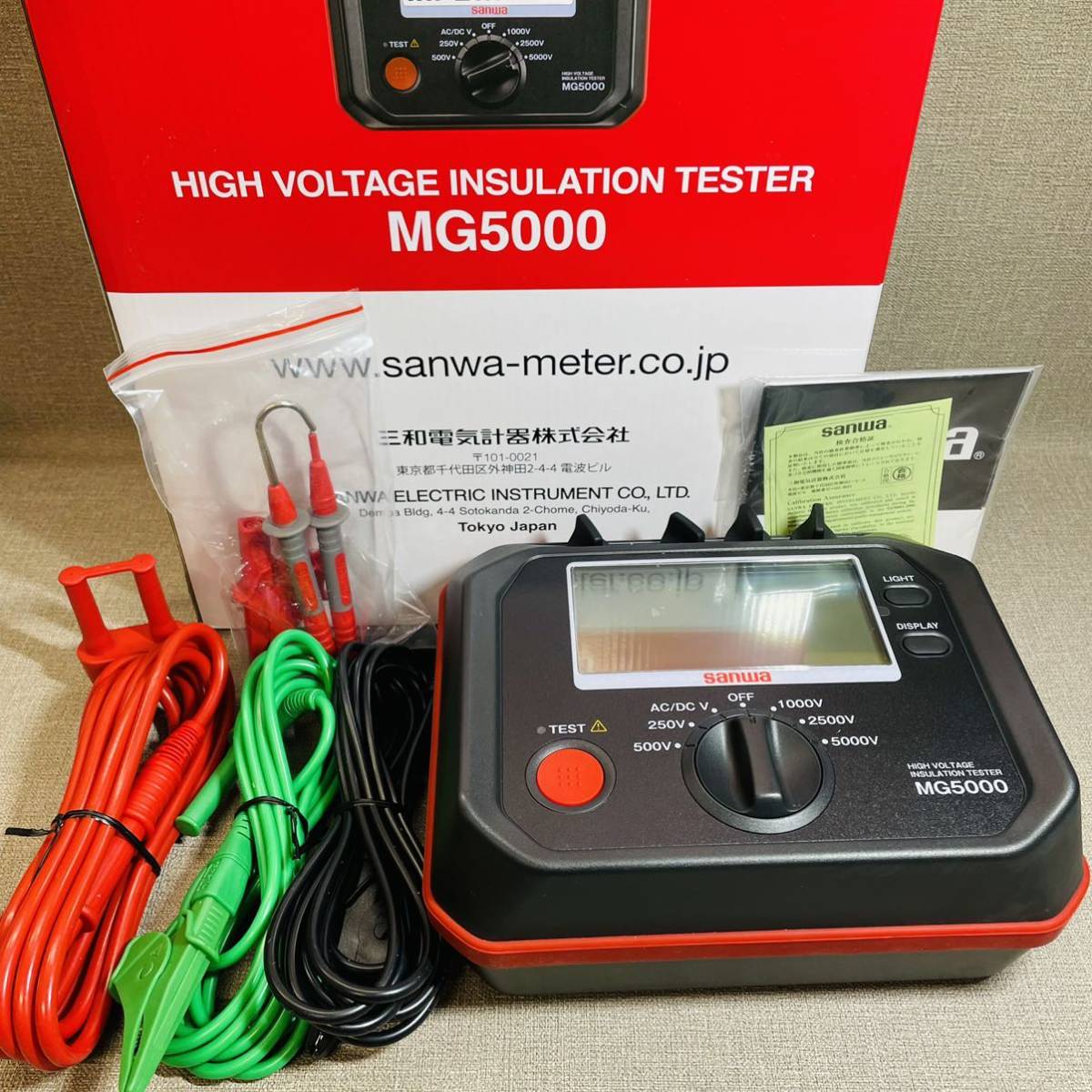 Sanwa MG5000 insulation tester powered off with test leads, clips, user manual, calibration certificate, and retail packaging beside the device.