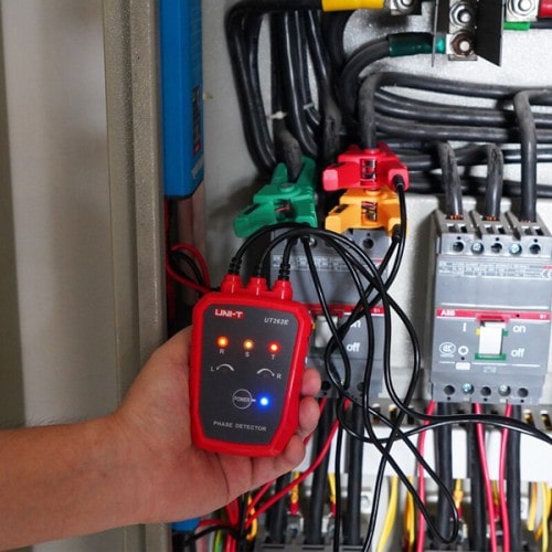 A technician connects leads to a motor control switch inside a control panel using the UNI-T UT262E phase rotation tester, LED indicators show phase sequence.