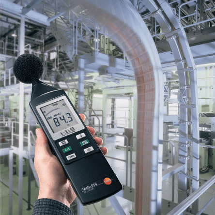 A technician using the testo 815 sound meter to measure occupational noise levels in an industrial facility with HVAC ducts in the background.