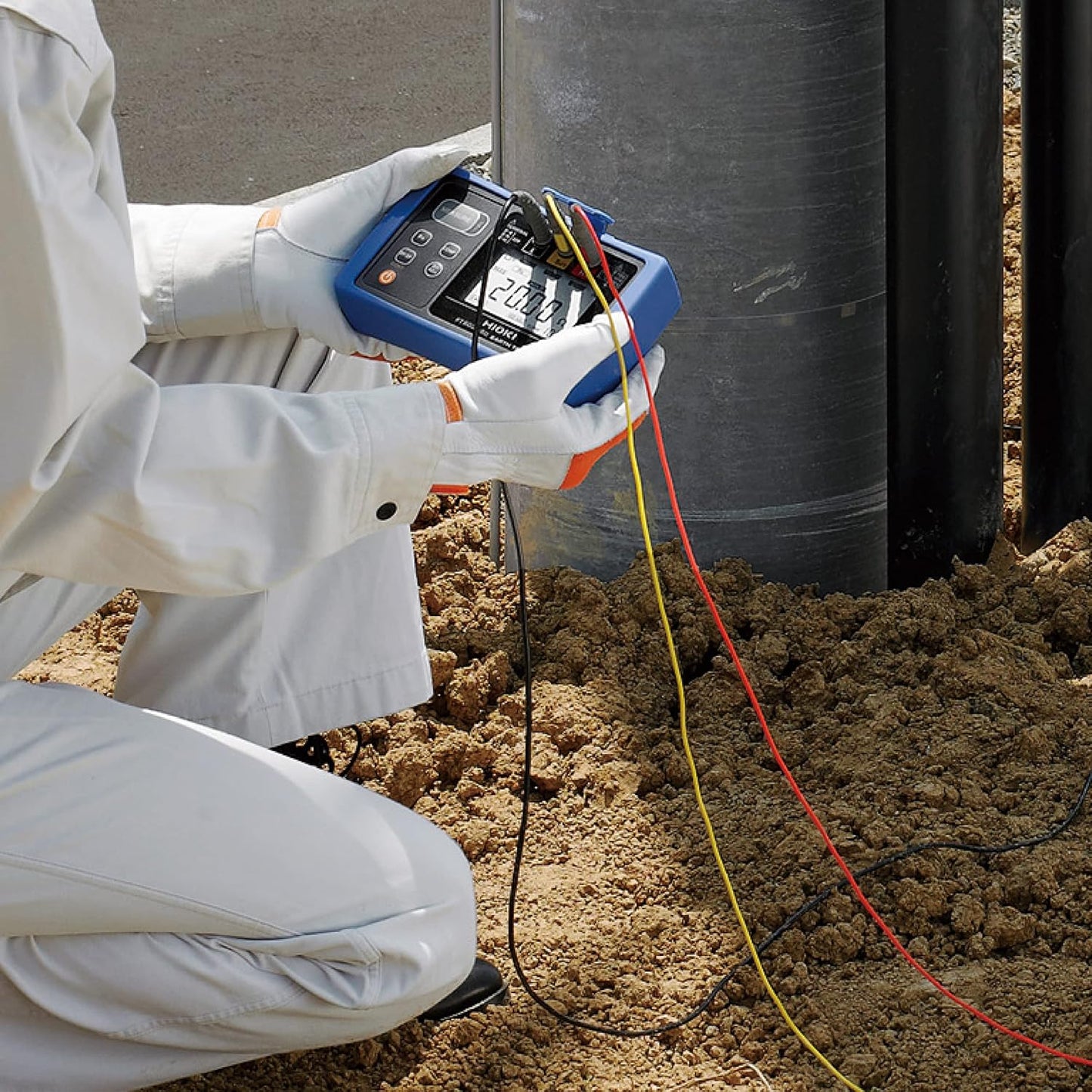 A technician performs an earth pit resistance test using the HIOKI FT6031-50, with test leads connected to the ground in sandy soil.