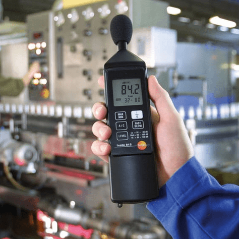 An industrial technician holding the testo 815 decibel meter to assess noise exposure near a machine control panel.