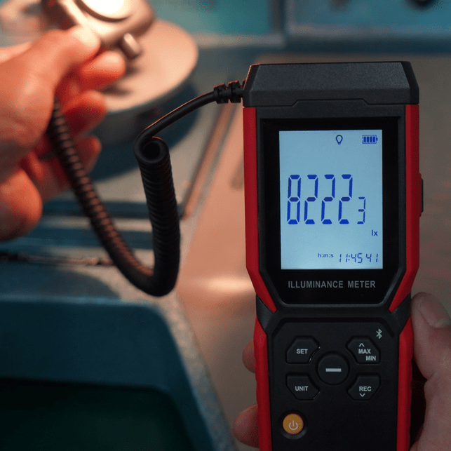 Technician holding UNI-T UT381A meter and detached probe to measure workshop light, displaying 8222.3 Lux on the screen.