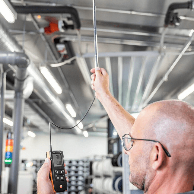 Technician using the TESTO 425 hot- wire anemometer to measure air flow around an overhead industrial pipeline, demonstrating its use in complex plant environments.