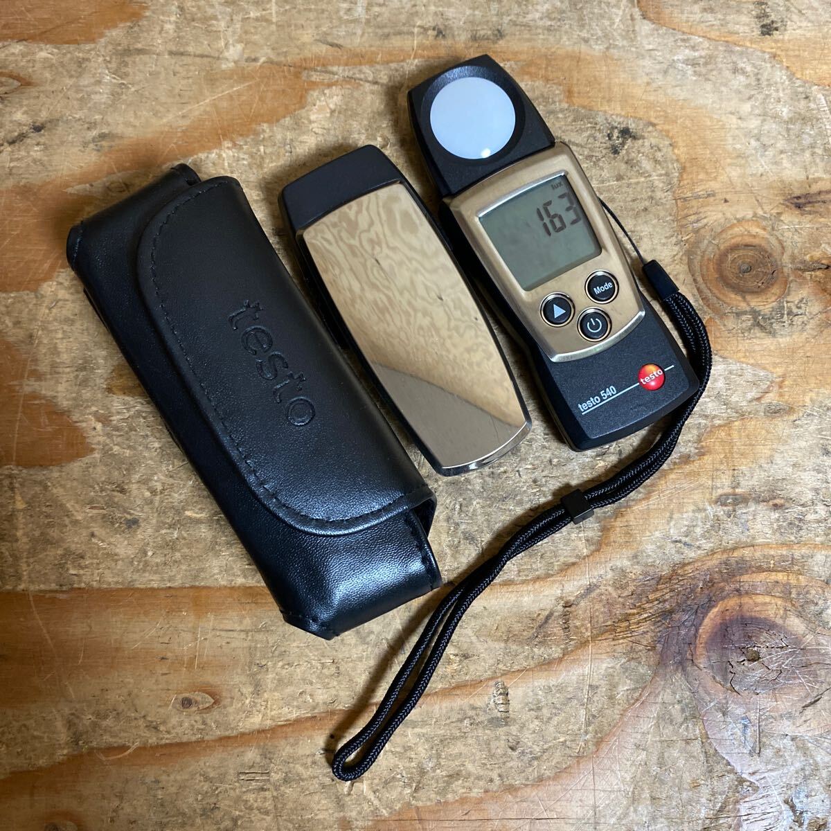 The TESTO 540 light meter with its protective cap removed, laid next to its belt pouch on a wooden surface, displaying a 163 Lux reading.
