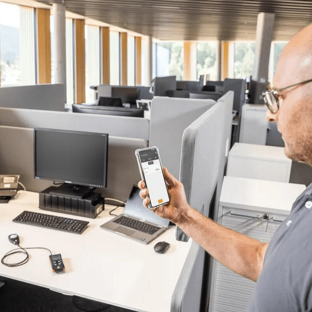 The Testo 545 digital lux meter operating on a desk in an office environment, with a technician monitoring real-time light measurement data via the Testo Smart App.
