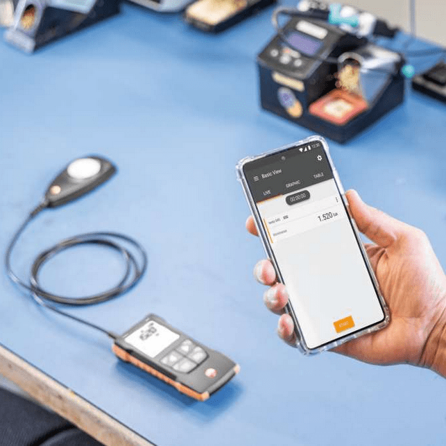 The Testo 545 lux meter in operation mode on a workbench beside calibration equipment, showing accurate light level readings on the display.