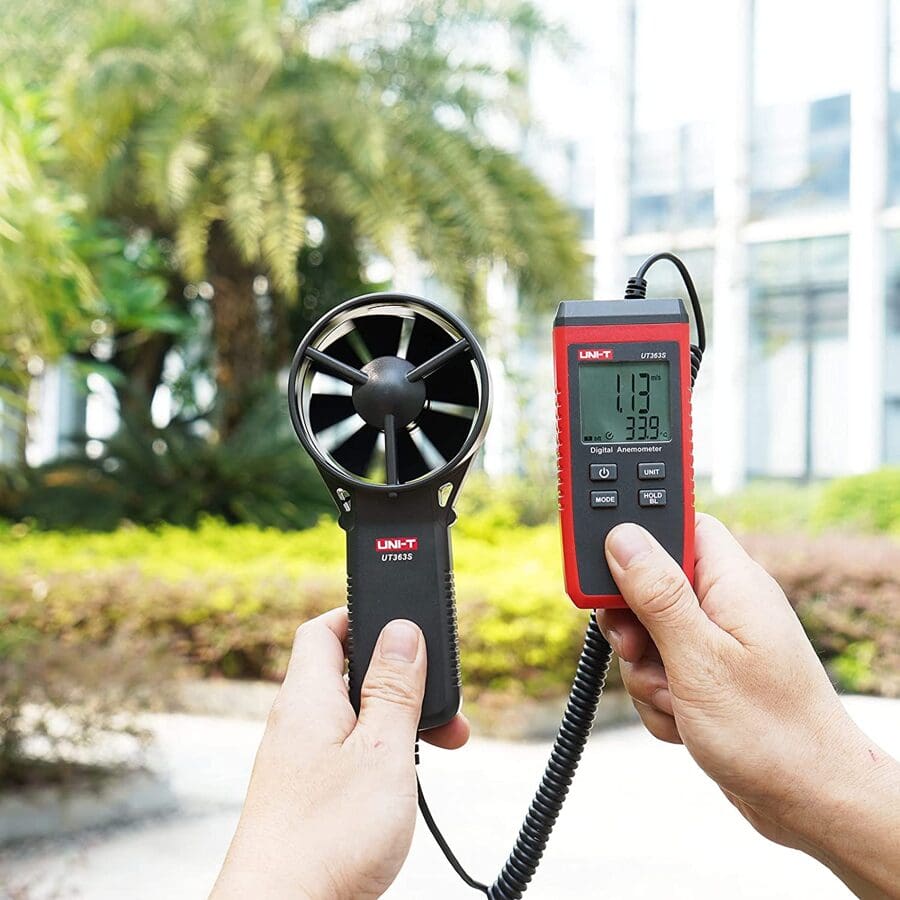 A technician uses the UNI-T UT363S handheld anemometer to measure outdoor wind speed, with the display showing 1.13 m/s in a lawn and garden setting.