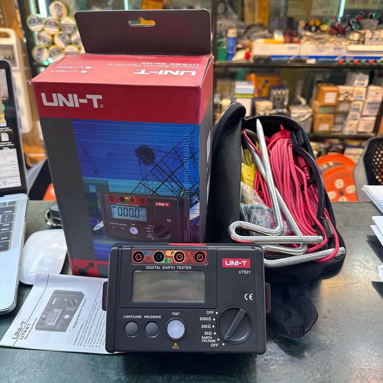 The UNI-T UT521 earth resistance meter kit laid out in a workshop, showing the handheld tester alongside its probes, spikes, and professional packaging.