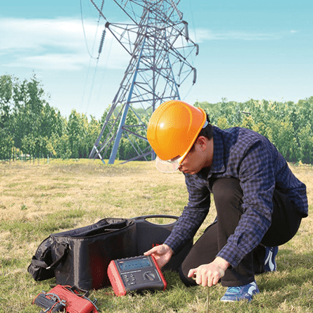 UNI-T UT572 Digital Ground Resistance Tester in field operation near high-voltage transmission towers, technician performing 4-pole soil resistivity measurement for power system grounding compliance