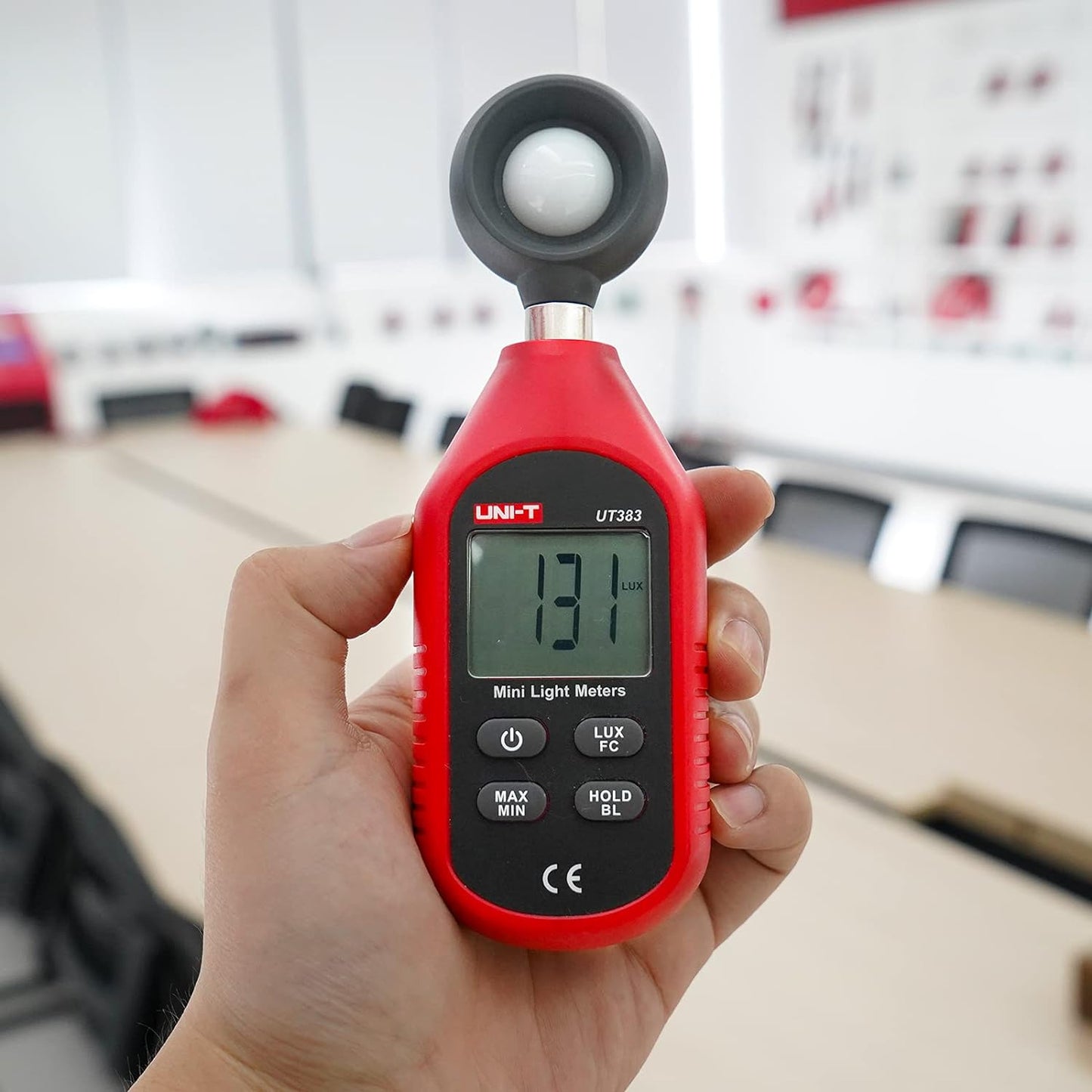 A person using the UNI-T UT383 digital light meter to check office lighting levels, with the device displaying a 131 Lux reading in a meeting room.